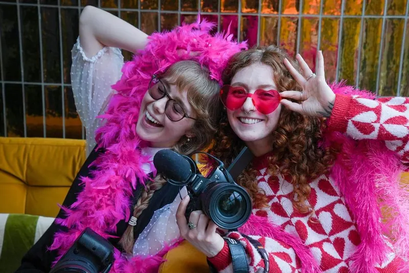 Two Women's Circle guests laughing together, one in a pink feather boa and the other wearing heart shaped sunglasses