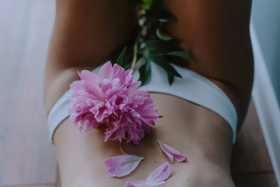 A soft, body positive close up of a pink peony flower resting on a woman's shoulder, with petals scattered below, a lifestyle image from Bang On Period
