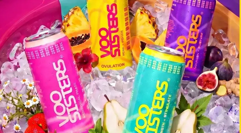 The four VOO Sisters cycle phase drinks: pink, yellow, purple, and teal cans chilling on ice, surrounded by fresh fruit and flowers