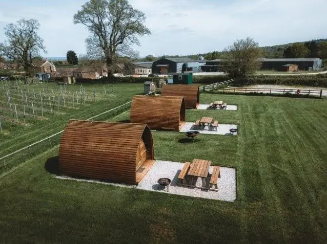 Aerial view of three wooden glamping pods with private fire pits and picnic benches, set beside a vineyard