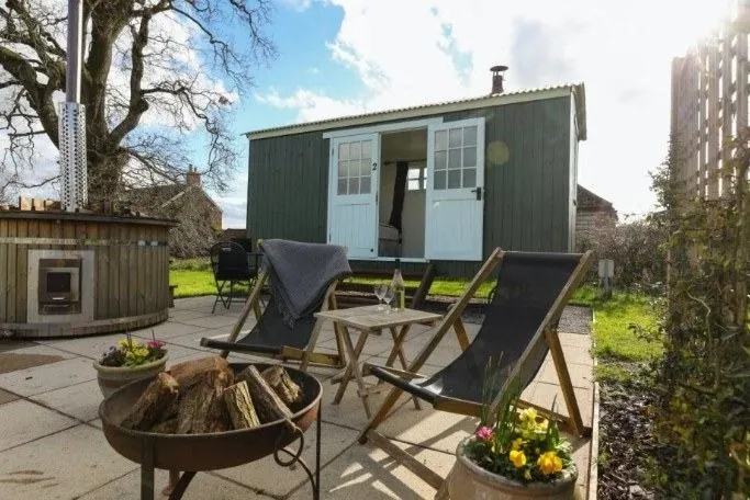 A cosy shepherd's hut with a wood fired hot tub, deck chairs and a fire pit on the patio